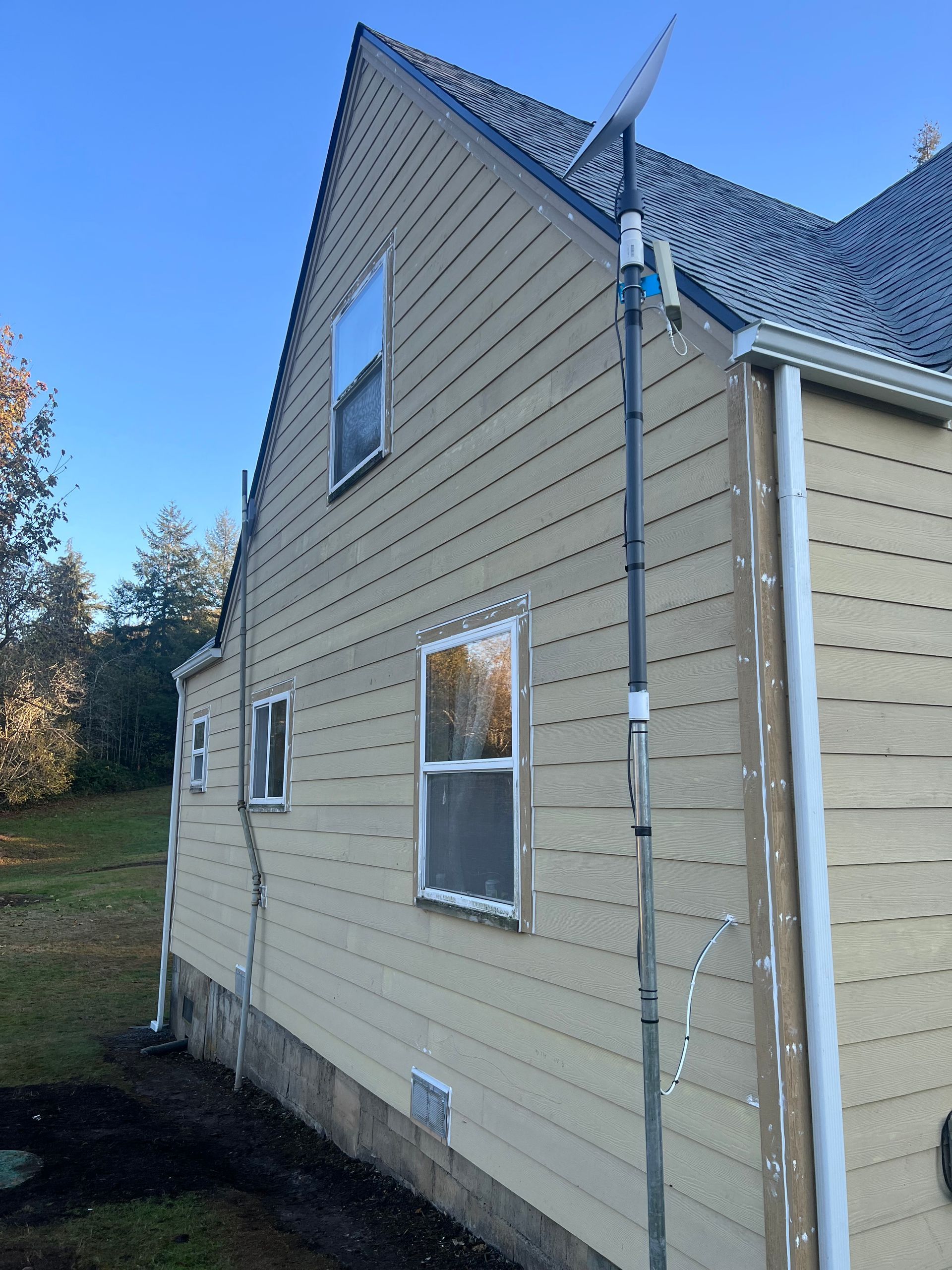 A tan-sided house with a Starlink satellite dish mounted on a vertical pole on the right and an electrical pipe on the left.