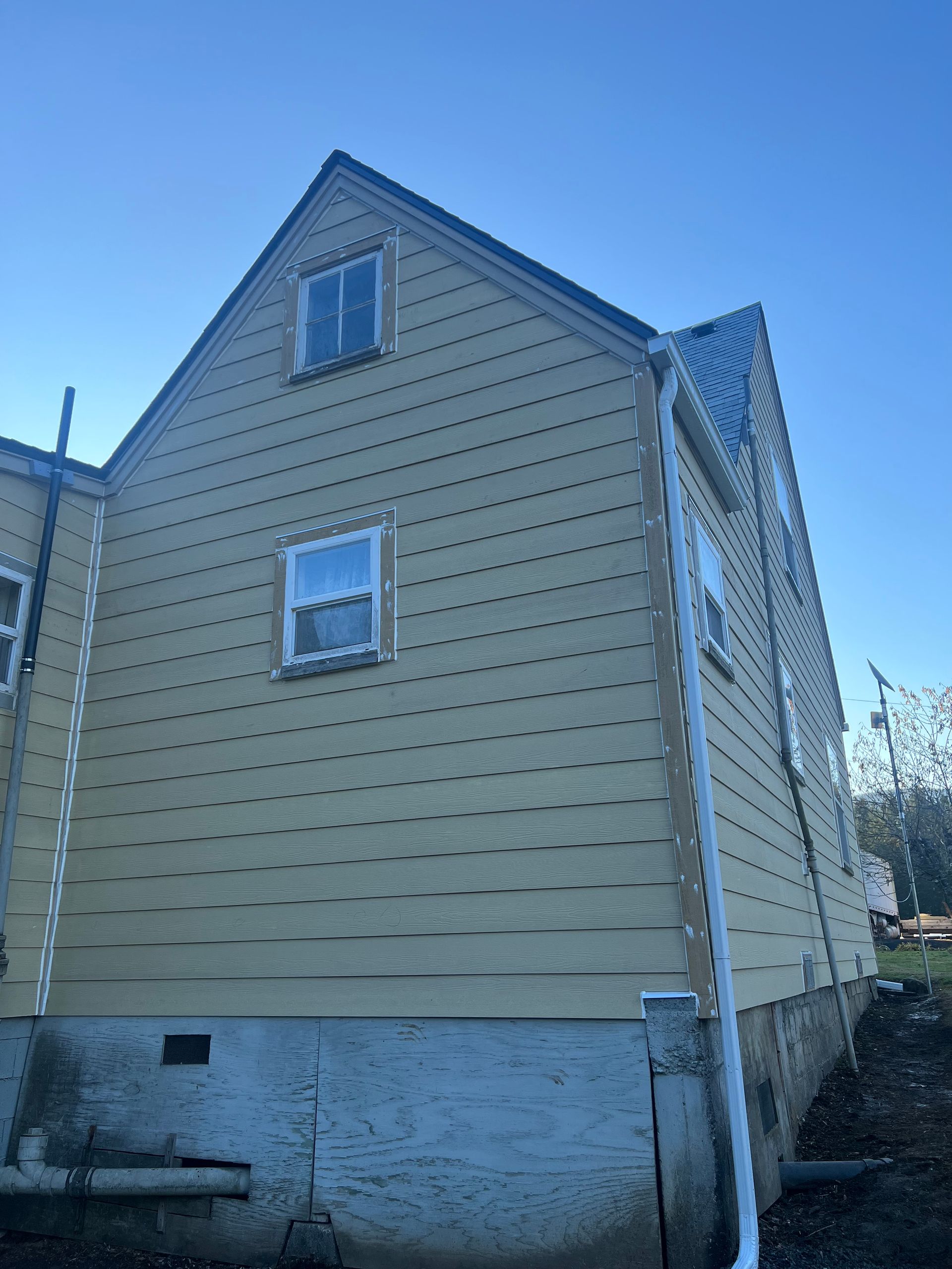 A yellow, wood-sided house exterior with a grey concrete foundation, two windows, and a white gutter under a blue sky.