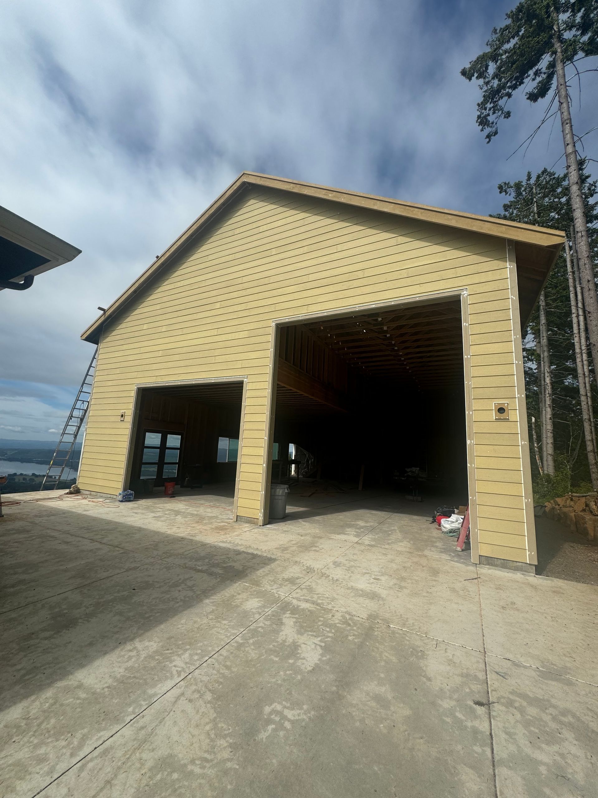 A tan building under construction with two large open garage bays, set on a gravel area under a cloudy sky.