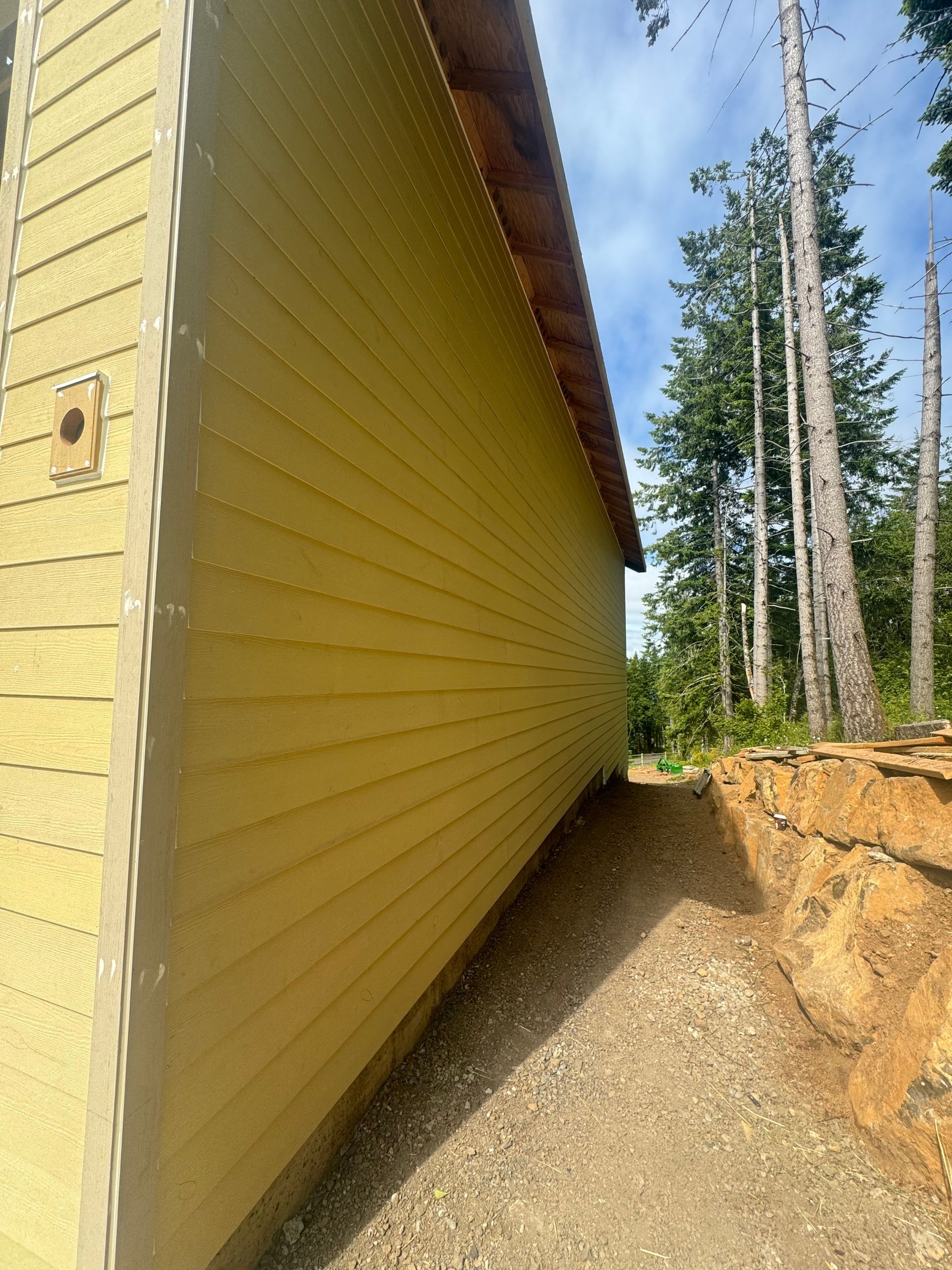 Yellow siding on the side of a building next to a dirt path and a rock retaining wall, with trees in the background.