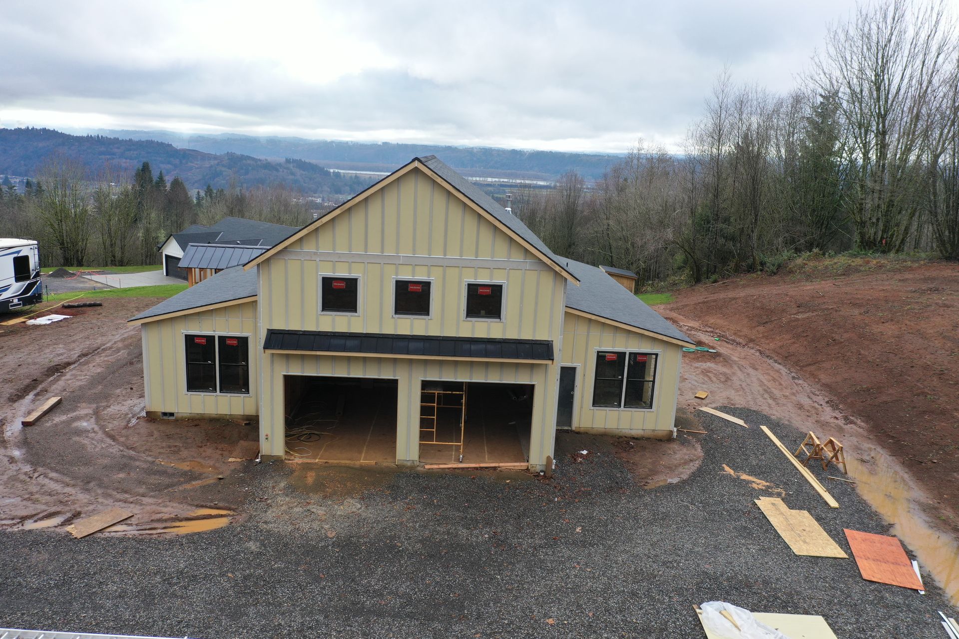 An exterior view of a new, beige, barn-style house under construction with a gravel driveway and hilly, forested landscape.
