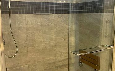 Shower stall with gray tile walls, glass door, and a wood seat.
