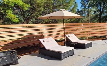 Two lounge chairs under an umbrella beside a pool, with a wooden fence in the background.