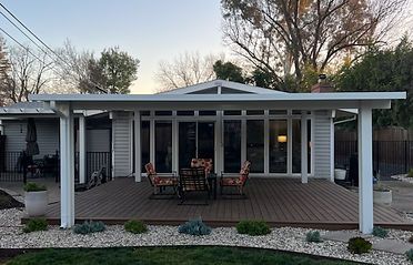 Backyard deck with patio furniture under a white roof, facing a white house, surrounded by gravel and greenery.