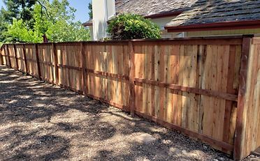 Wooden fence in a yard, brown with dark support beams, in front of a house with trees.