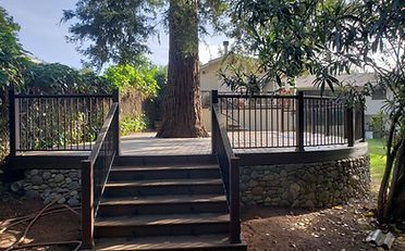 Wooden deck with steps, black railing, built around a large tree in a yard.