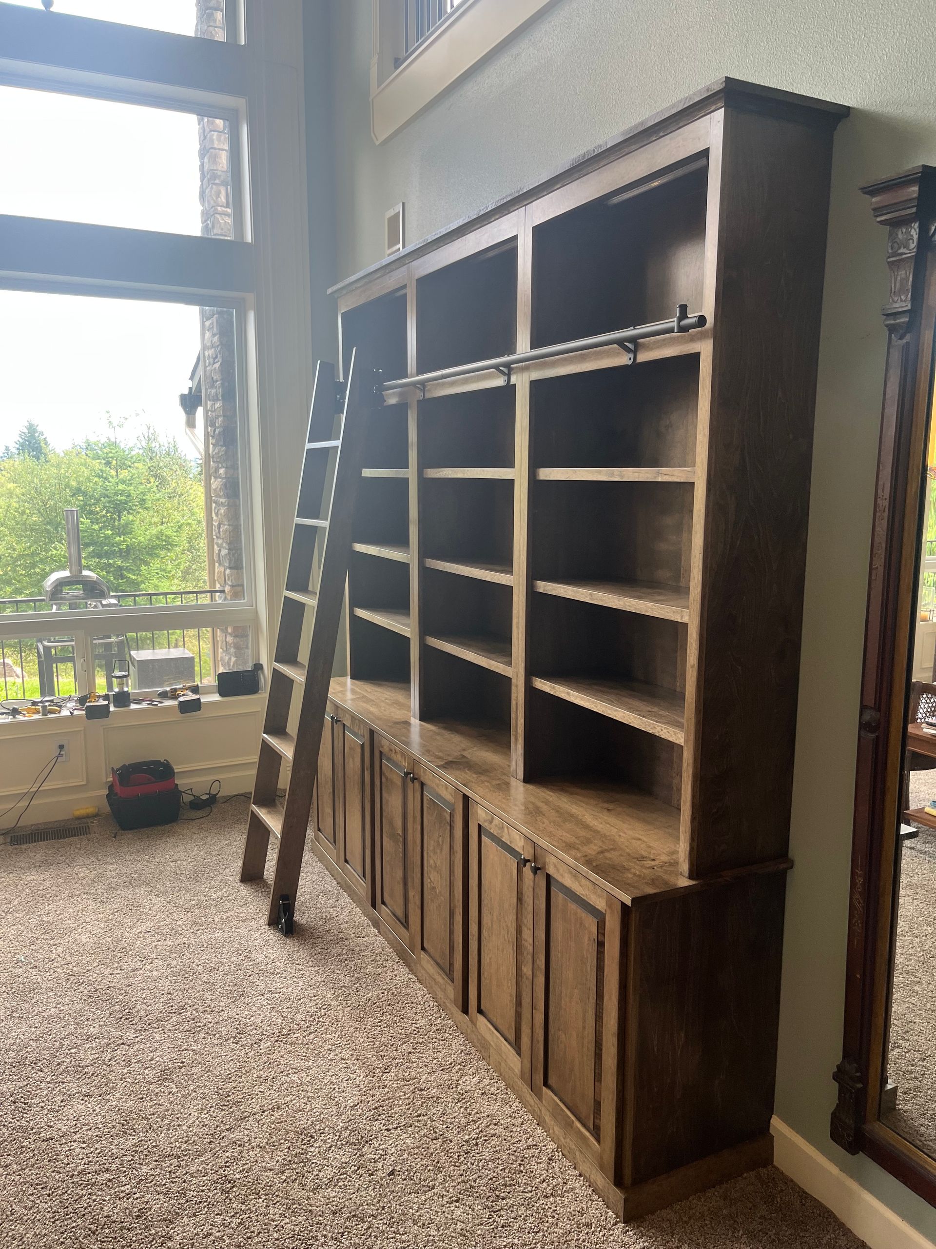 Tall wooden bookcase with a ladder in a room with a large window and carpet.