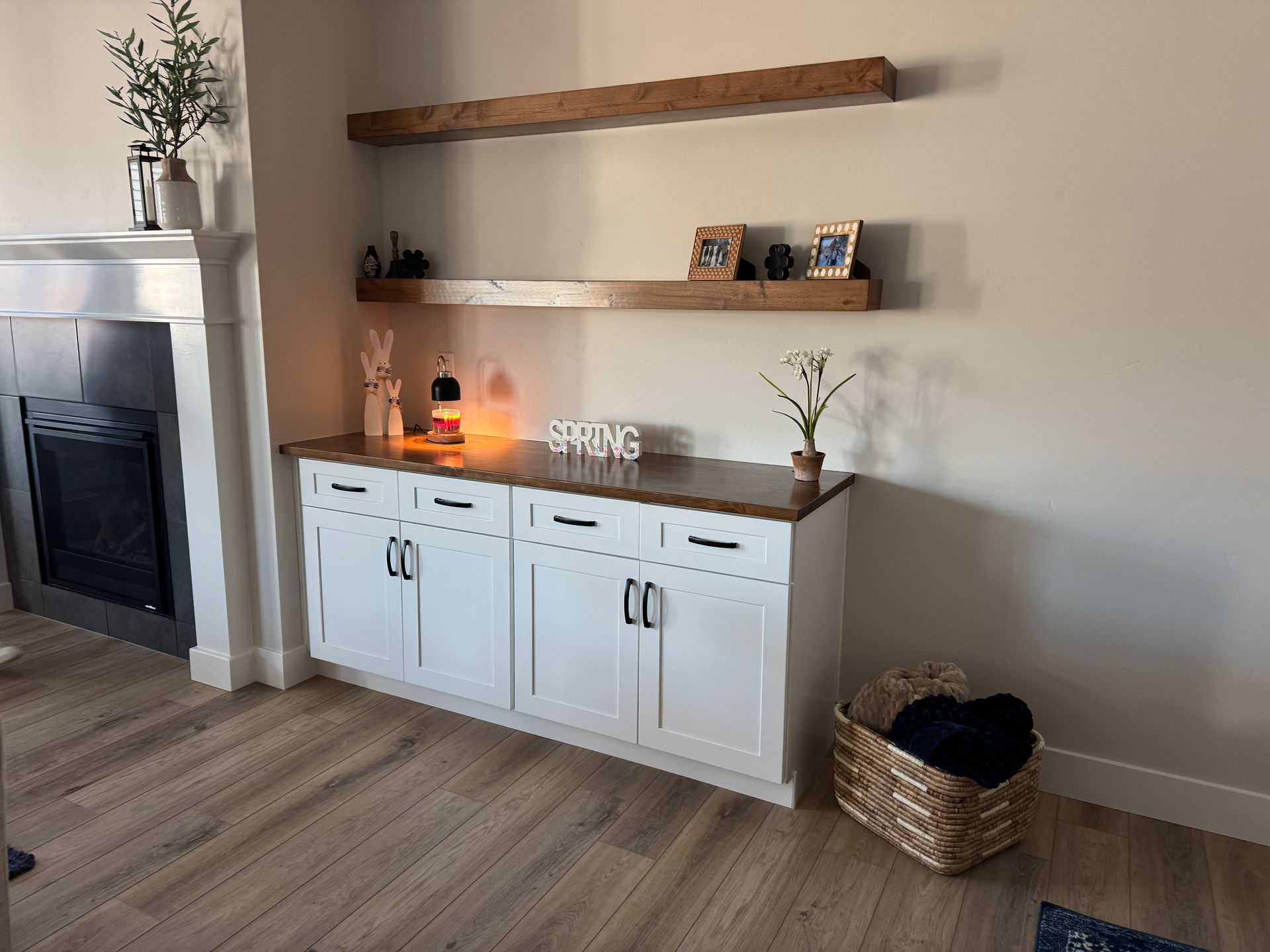 White cabinet with wooden shelves above, holding decor in a neutral-toned living room with fireplace.