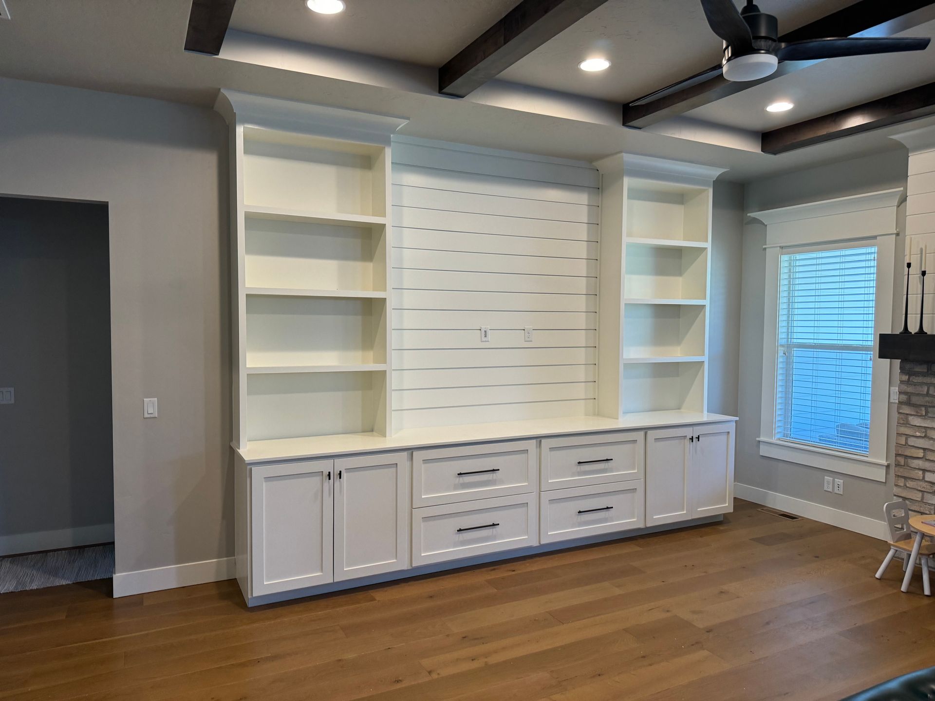 Built-in white entertainment center with shelving and storage cabinets in a room with wood flooring and a window.