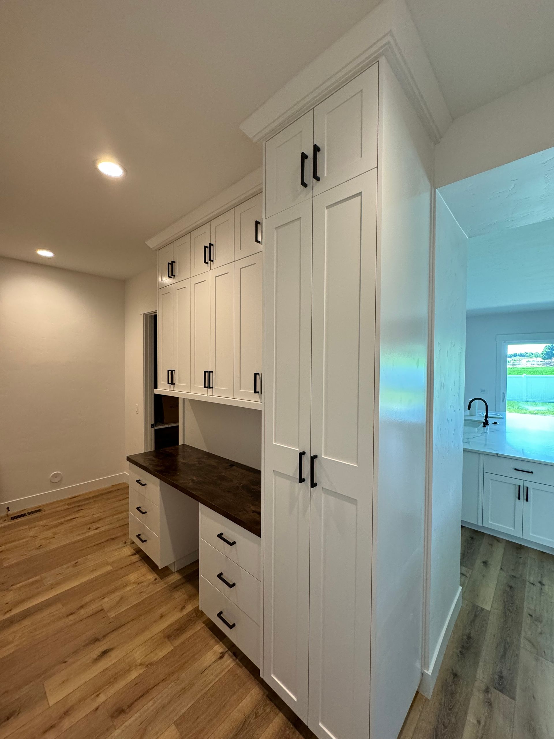 White cabinets with black hardware, a desk, and hardwood floors. View into a kitchen.