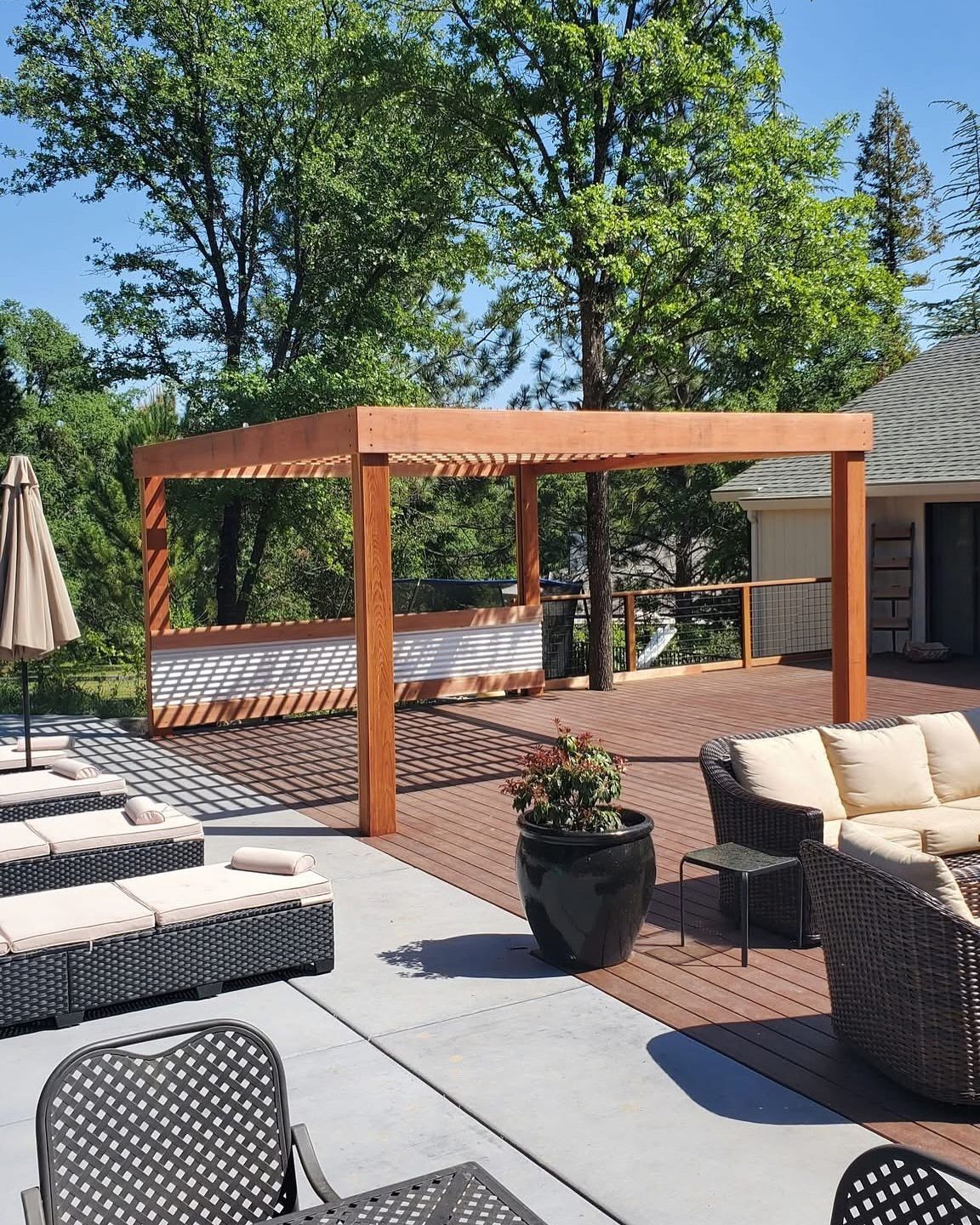 Wooden pergola on a wood deck, surrounded by patio furniture and sunbathers; trees and blue sky in the background.