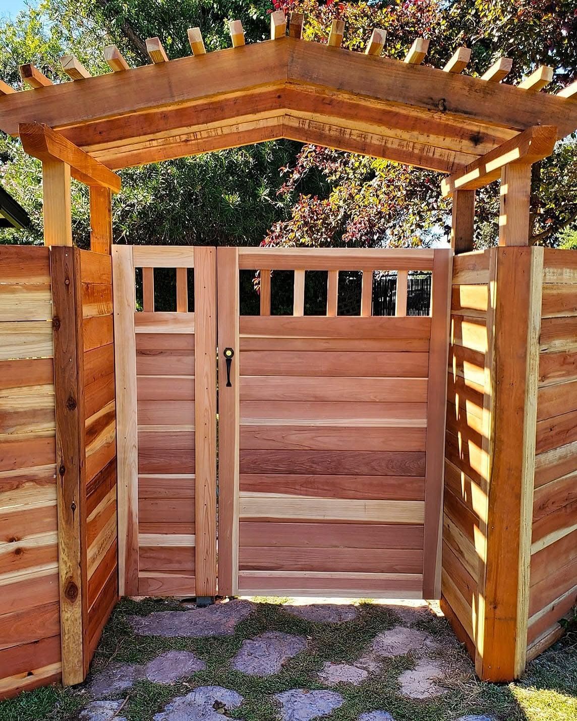 Wooden gate with pergola, surrounded by fence, on a stone path.