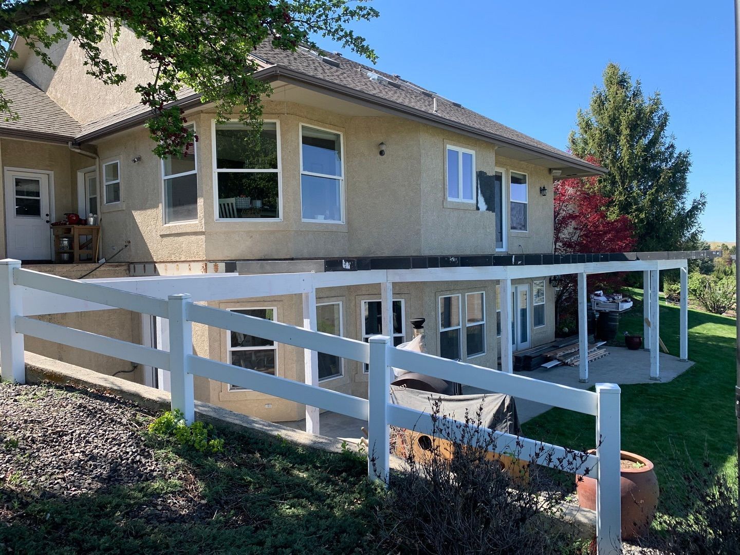 Beige house with white fence, lower level patio, and a green lawn under a blue sky.