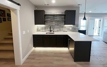 Dark gray wet bar with white countertop, cabinets, backsplash, and open shelving in a modern home.