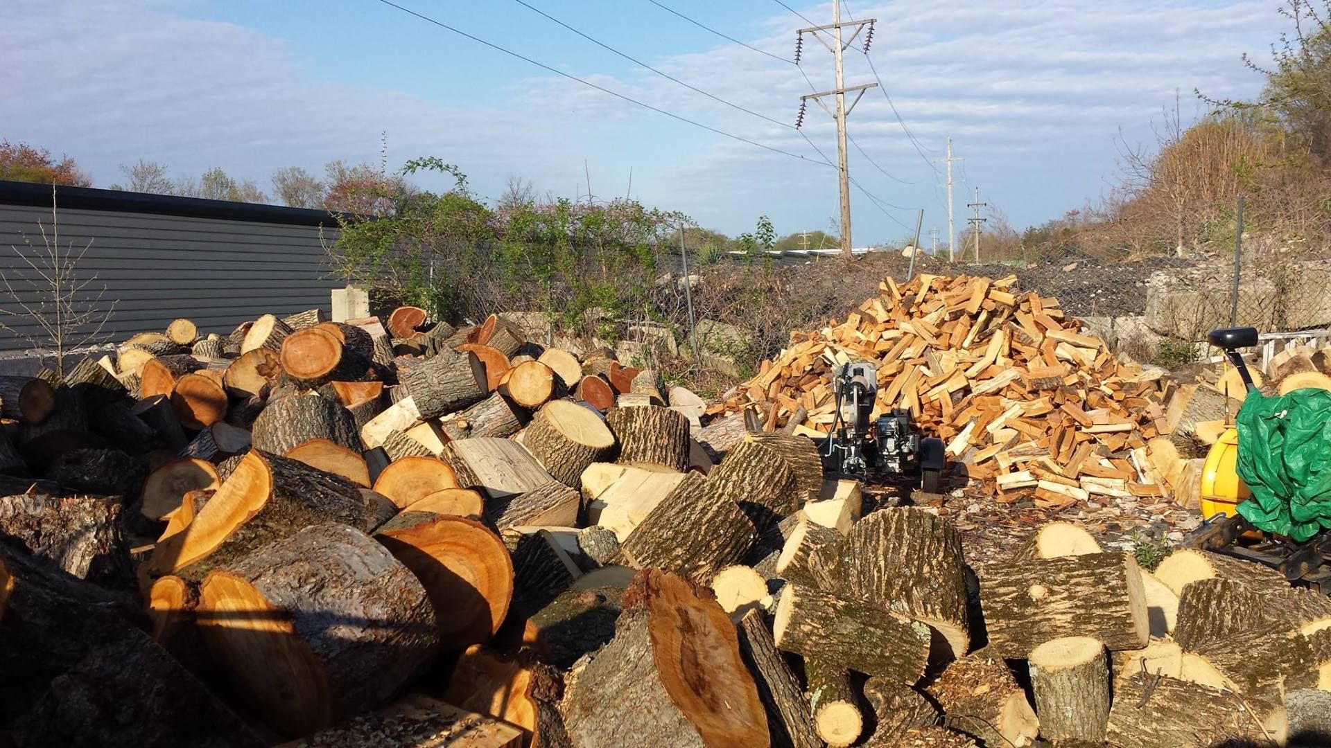 Firewood piles stacked outdoors beside a rural road, with power lines and a clear sky.