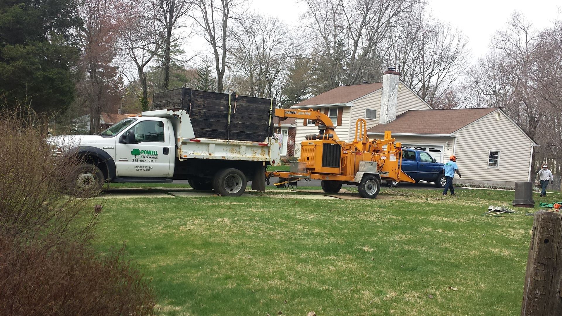 White utility truck, orange wood chipper, and blue car in a yard beside a house.