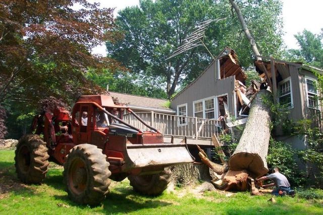 Bulldozer lifting a fallen tree beside a house while a person stands nearby in a yard