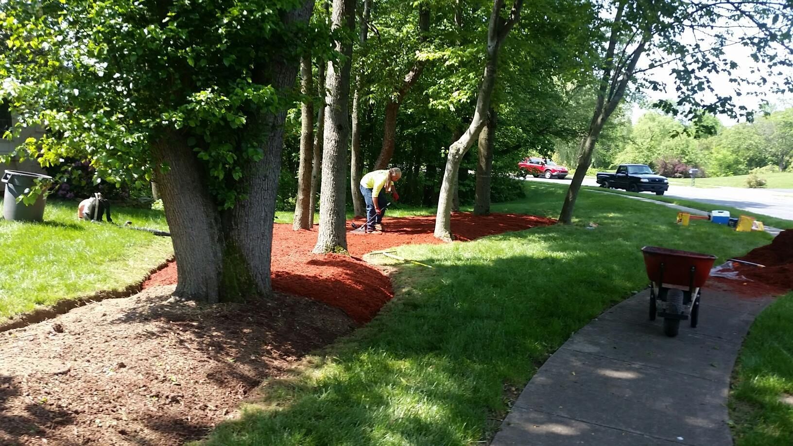 Shaded park walkway beside trees, red mulch beds, and a wheelbarrow on the path