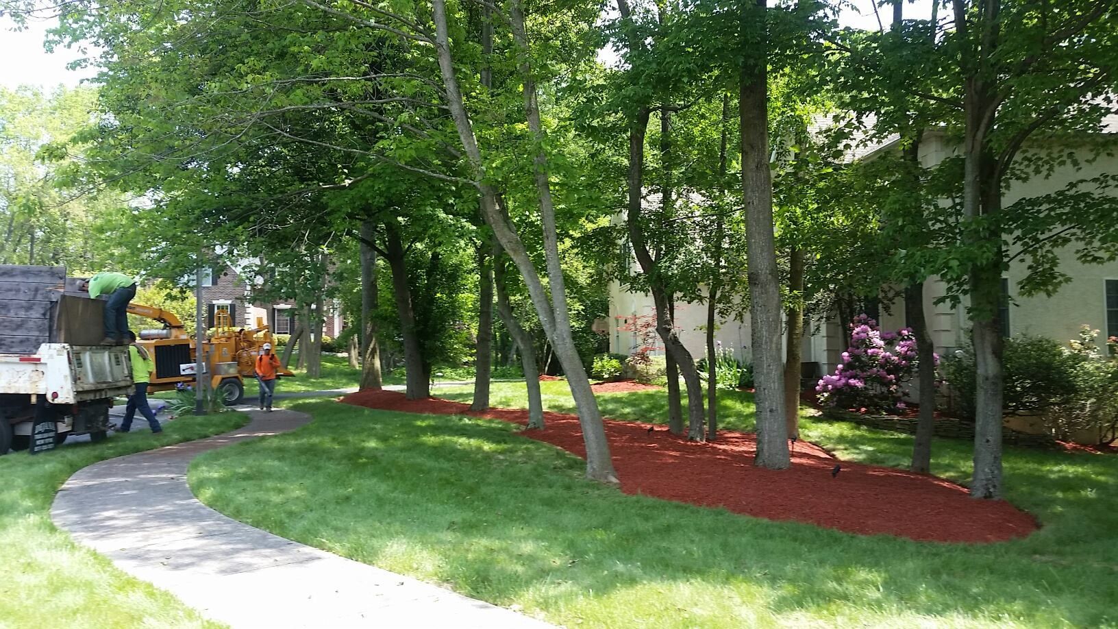 Park with curved sidewalk, trees, red mulch beds, and workers near a truck on the left.