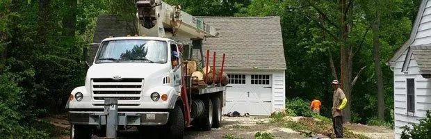 White utility truck with lift equipment parked by a house in a wooded yard; worker near the driveway