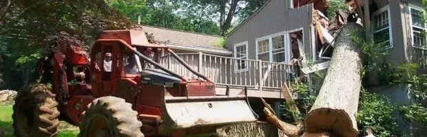 Red tractor and log loader lifting a large log in a wooded yard beside a house