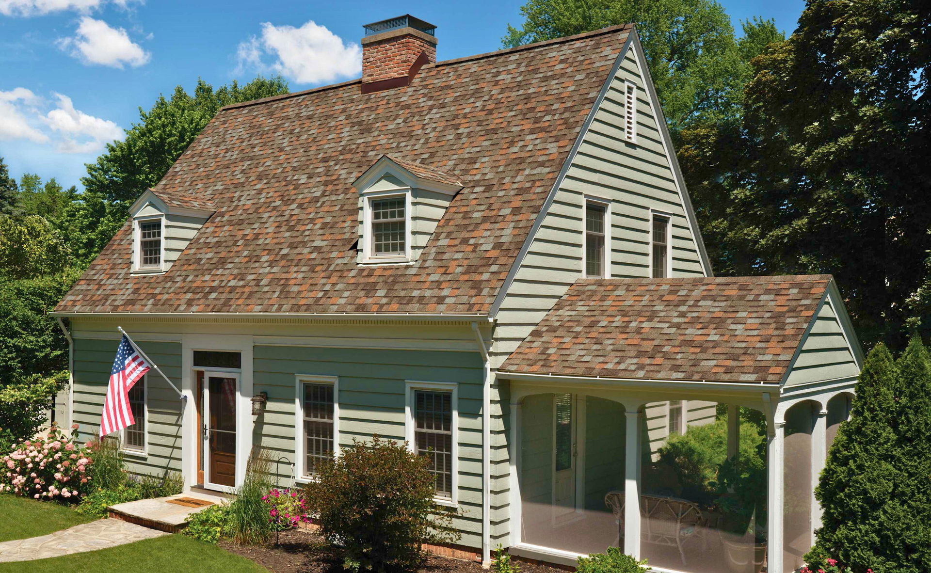 A light green two-story house with a brown shingle roof, American flag, and porch.