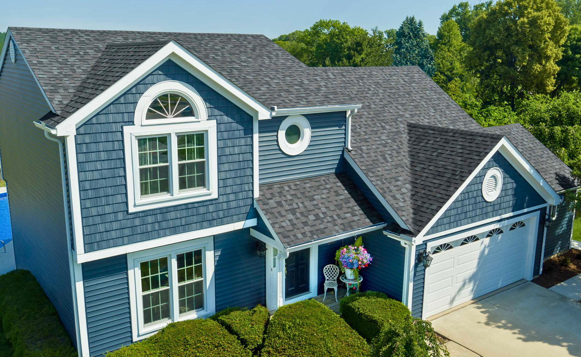 Blue house with white trim, dark roof. Front door with flowers, green bushes, and a garage.