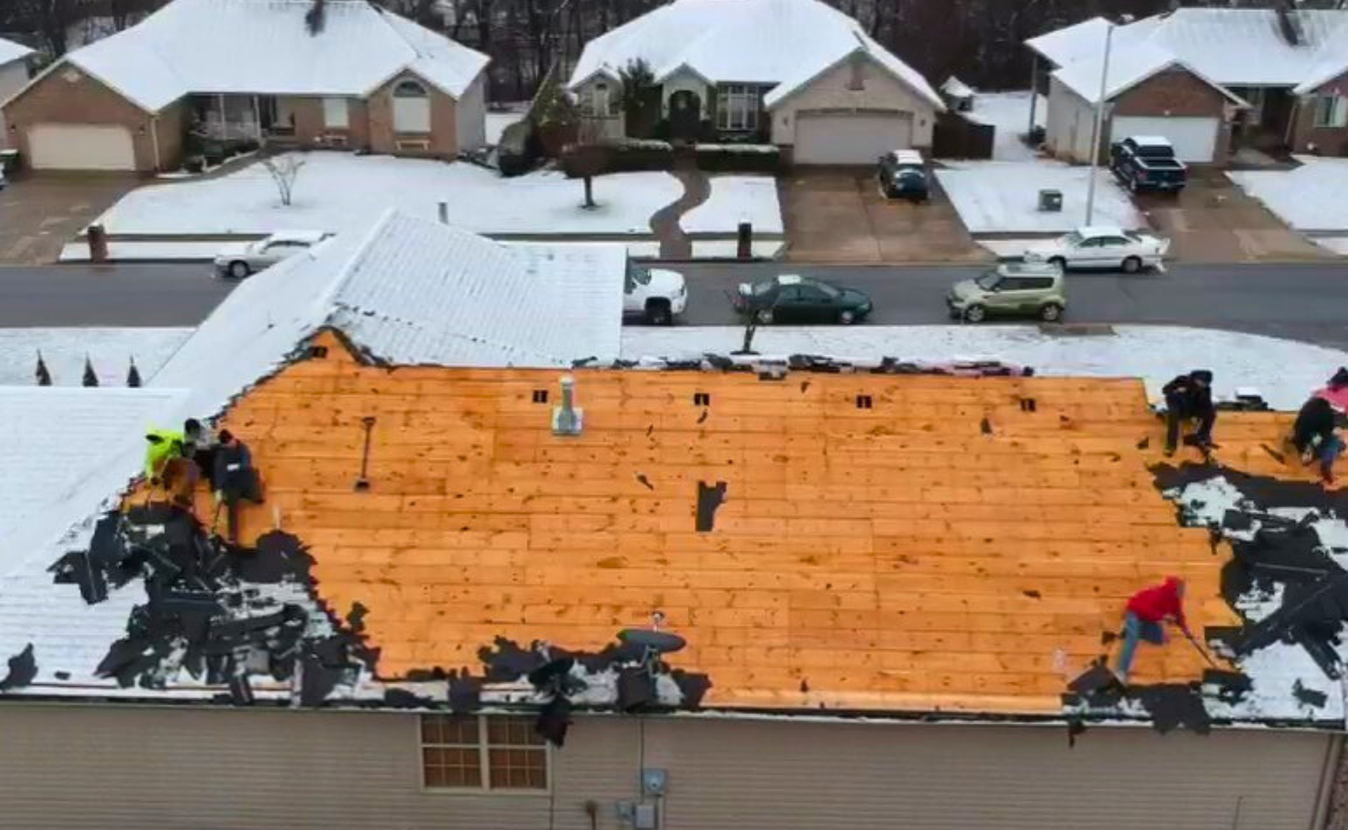 Roofers removing shingles from a house roof covered with snow in a residential neighborhood.