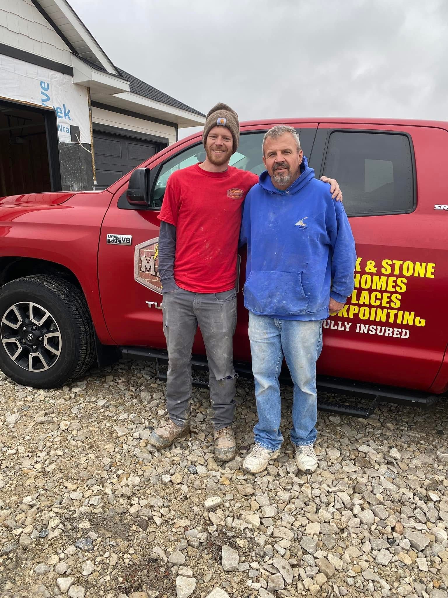 Two men stand by a red pickup truck. One wears a red shirt, the other a blue hoodie. Building in the background.