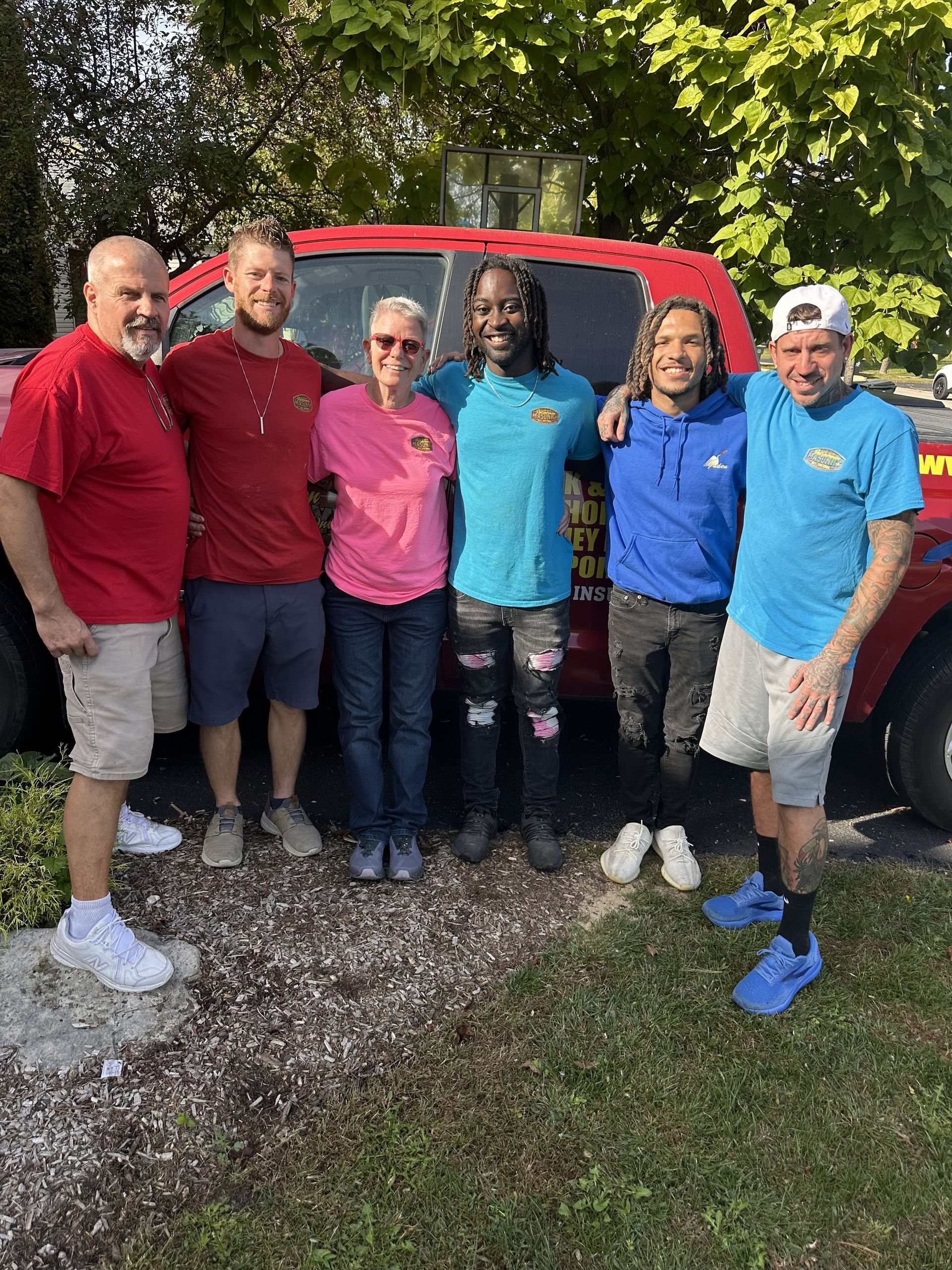 Group of six people posing together outdoors in front of a red truck; wearing various colored shirts.