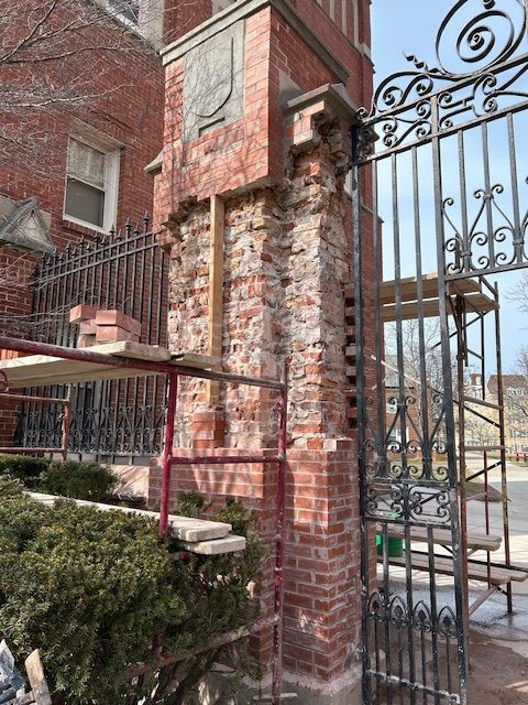 Brick pillar repair at ornate wrought iron gate; red brick, scaffolding, greenery.