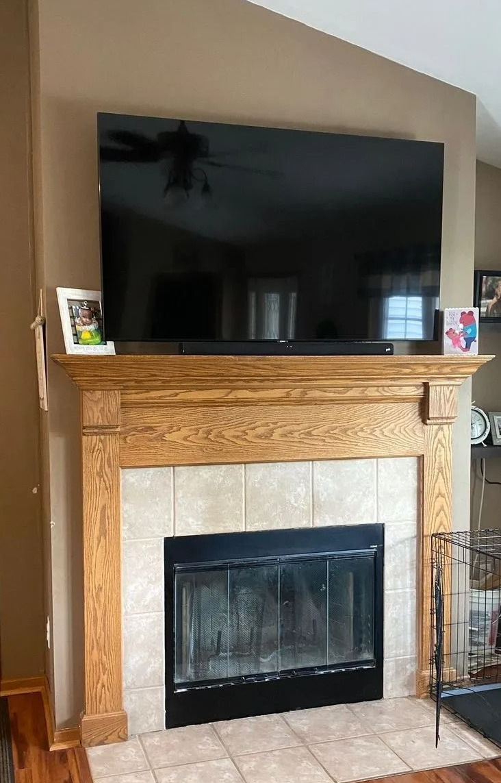 Living room with fireplace, TV above. Brown walls, wooden mantel, tiled hearth. Dark rug on wood floor.