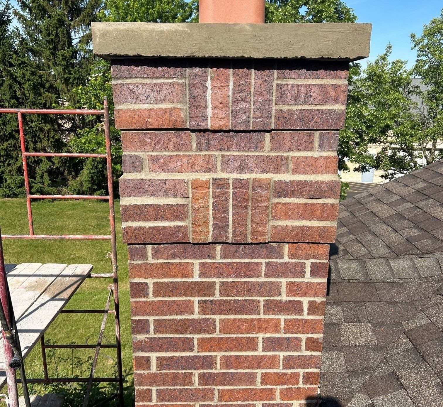 Brick chimney on a shingled rooftop with a red metal scaffold nearby and trees in the background