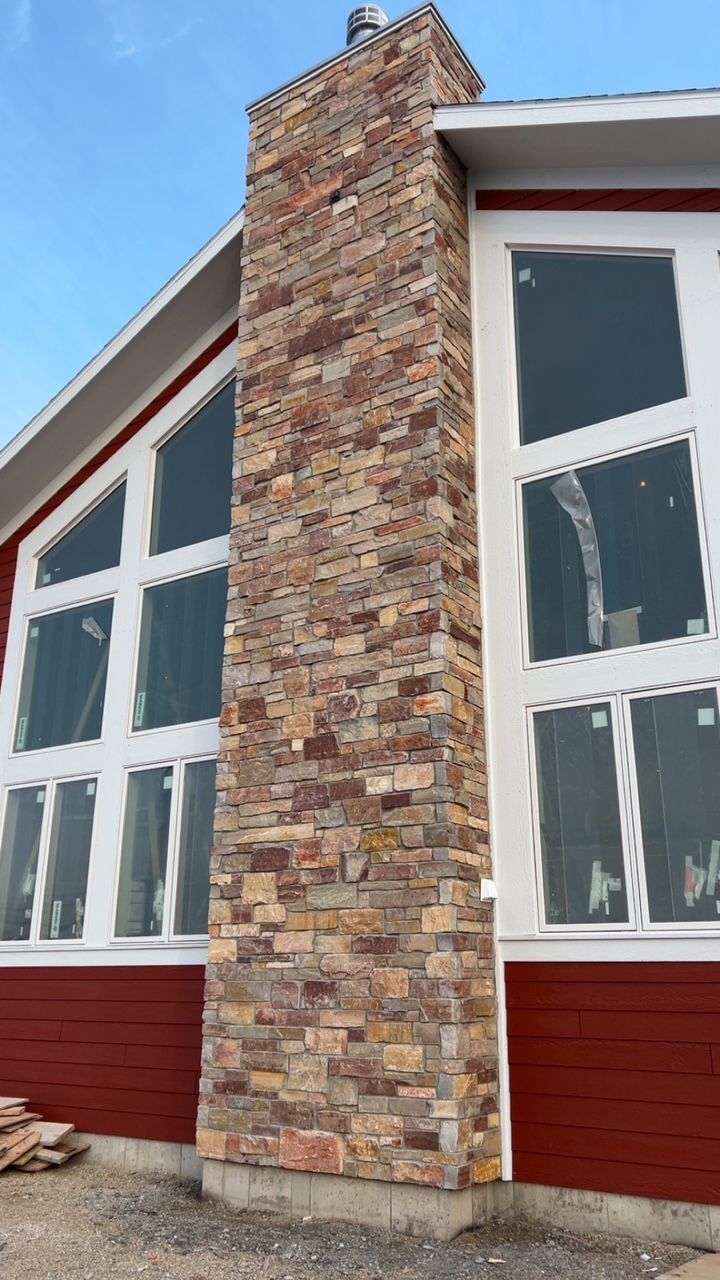A red-sided house features a tall, stone-faced chimney set between large, multi-pane windows against a clear blue sky.