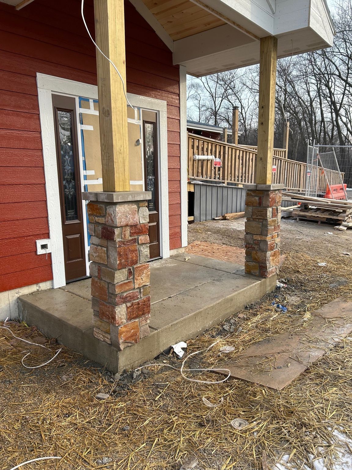 A partially constructed red building entrance with a concrete porch and two stone pillars supporting wooden porch posts.