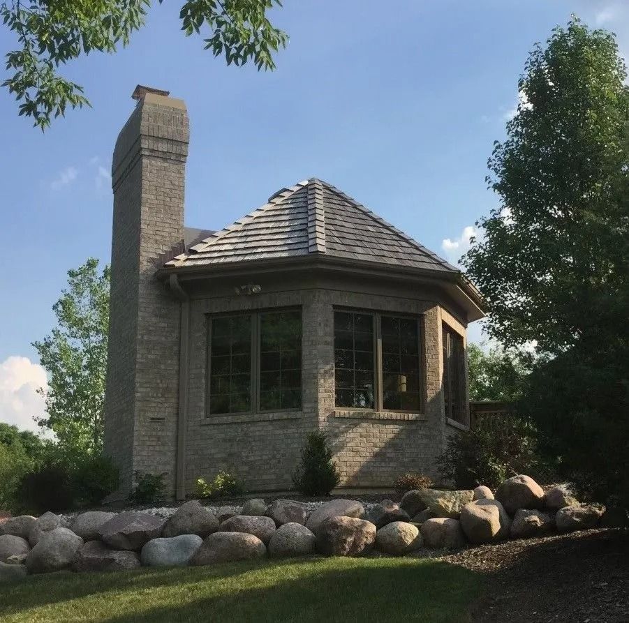 Brick building with a chimney, windows, and a shingle roof, set among rocks and trees.