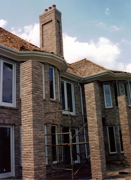 Brick house exterior with tall chimney, several windows, and scaffolding. Brown roof, blue sky.