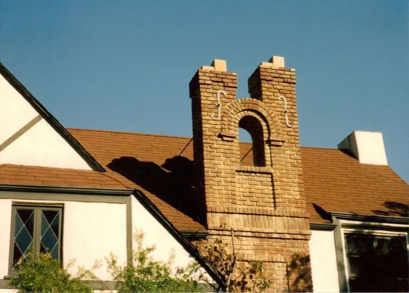 Brick chimney with arched opening atop a brown shingled roof against a blue sky.