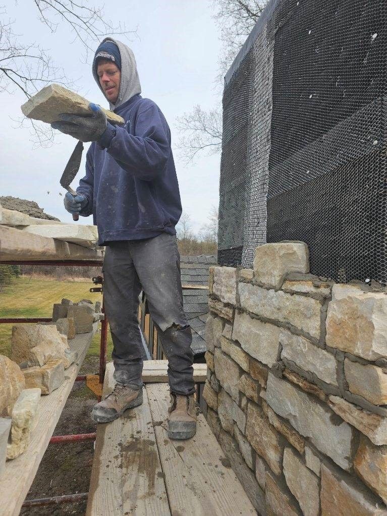 Man building a stone wall, holding a stone with a tool. He is standing on a wooden plank, working outdoors.