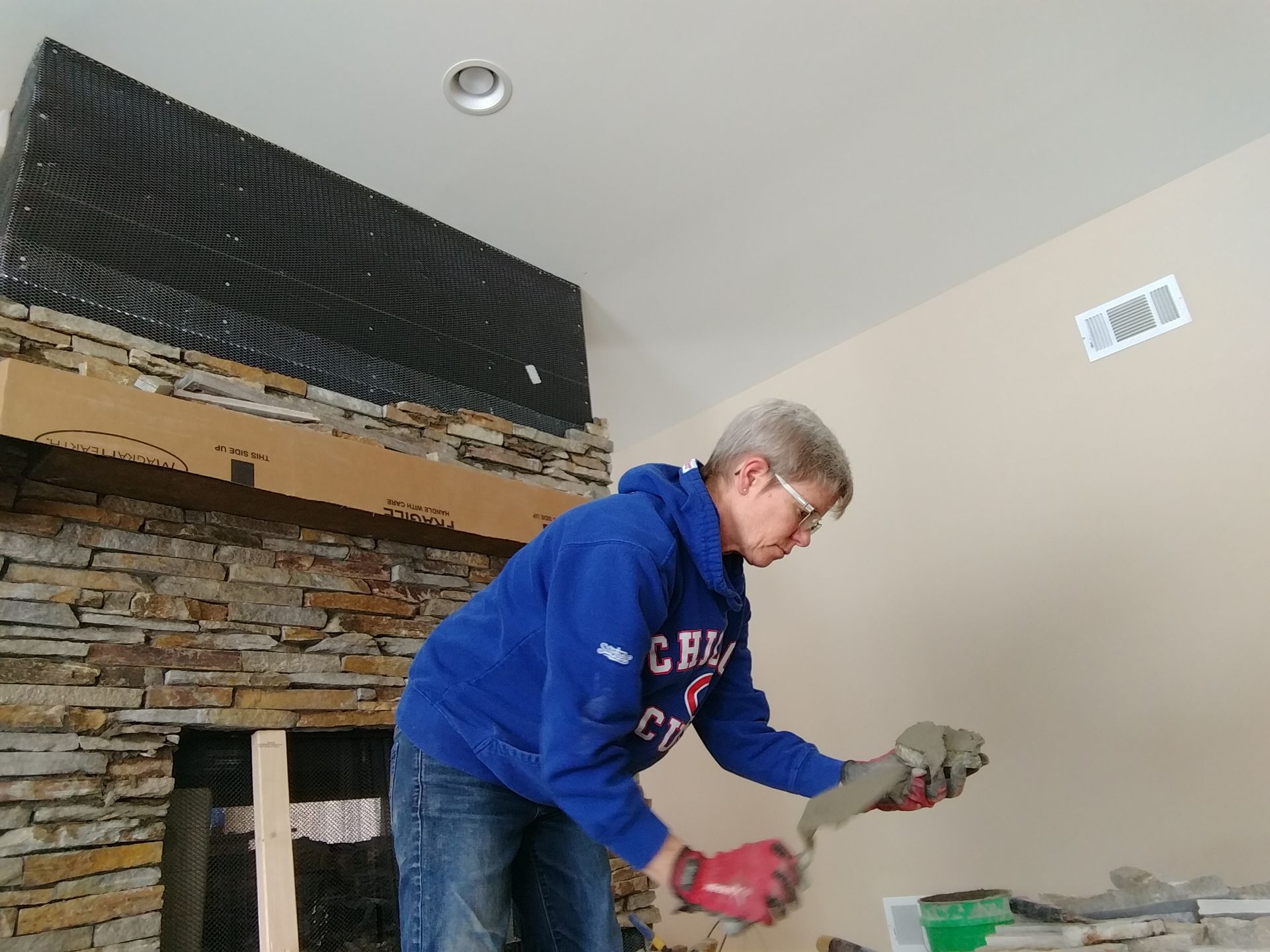 Person applying mortar to stones on a fireplace surround. Wearing blue hoodie, jeans, and red gloves.