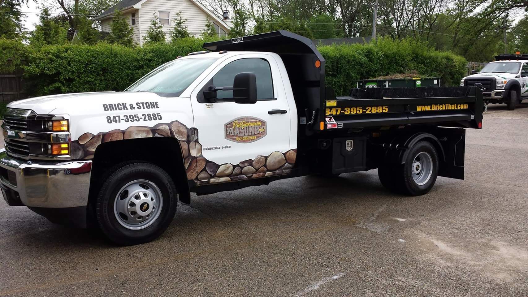 White dump truck with stone-patterned accents. Parked on asphalt with a green hedge background.