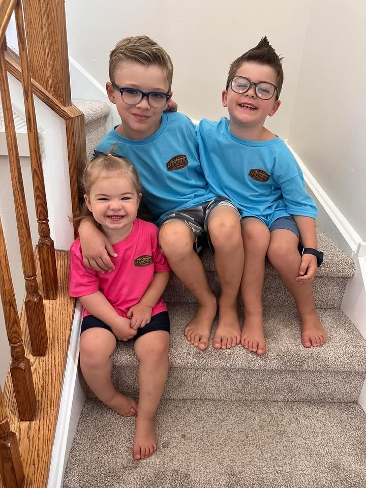 Three children smiling, sitting together on stairs, wearing colorful shirts and eyeglasses.