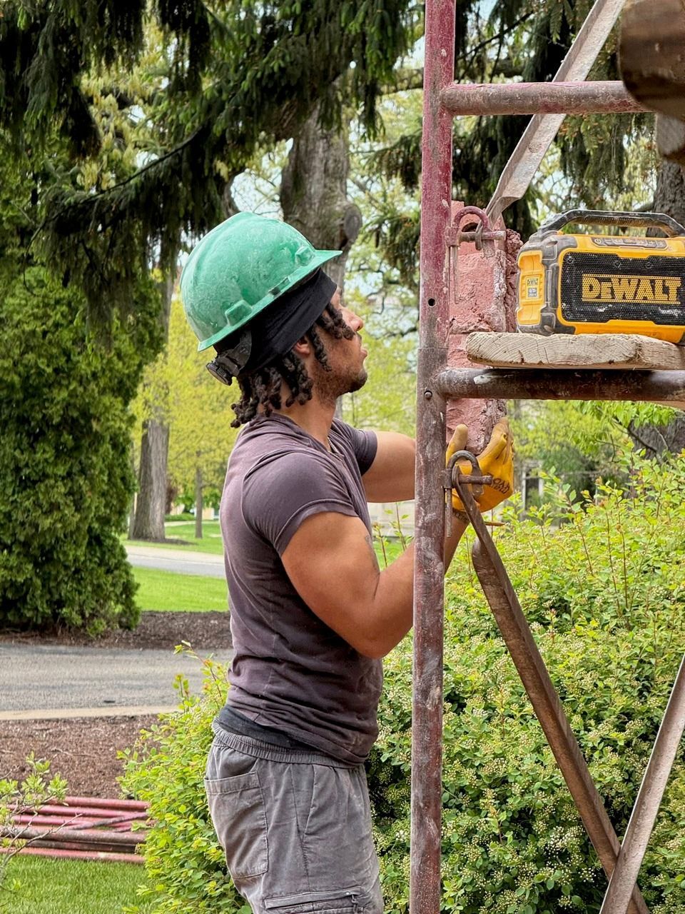 Construction worker in green hard hat on scaffolding, using a tool outdoors.