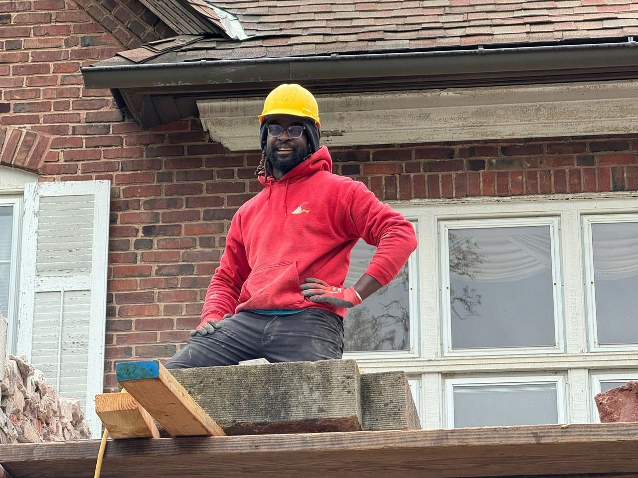 Construction worker wearing a yellow hard hat and red hoodie on a scaffold, in front of a brick building.