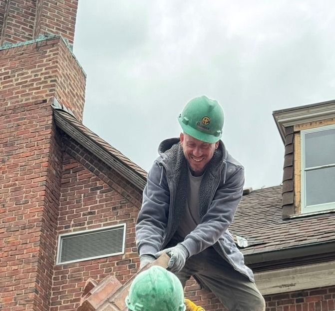 Man in hard hat working on a brick chimney, smiling. Cloudy sky, building in the background.