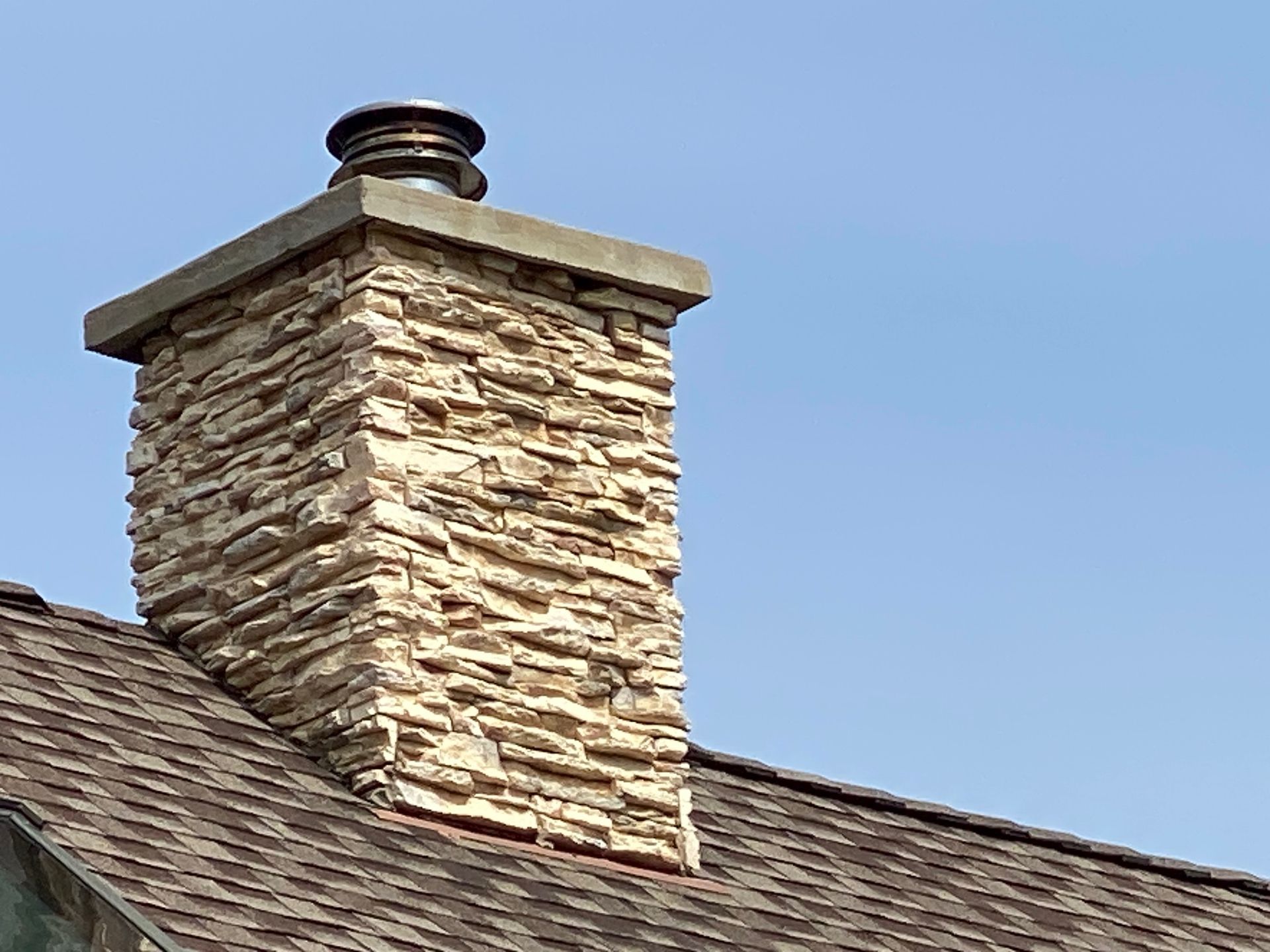 Stone chimney on a brown shingled roof against a blue sky.