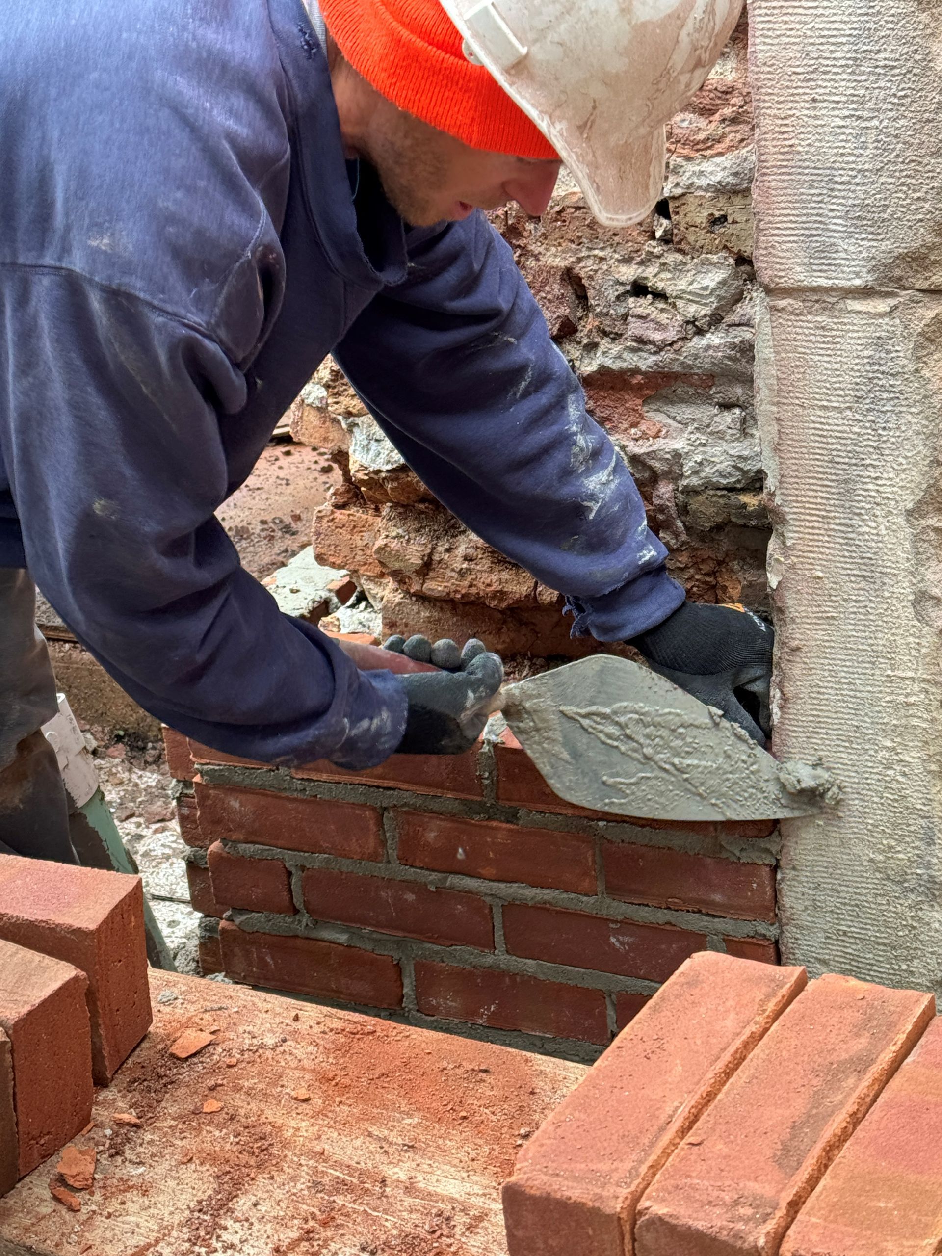 Construction worker laying bricks with a trowel at a building site; red bricks and mortar are visible.