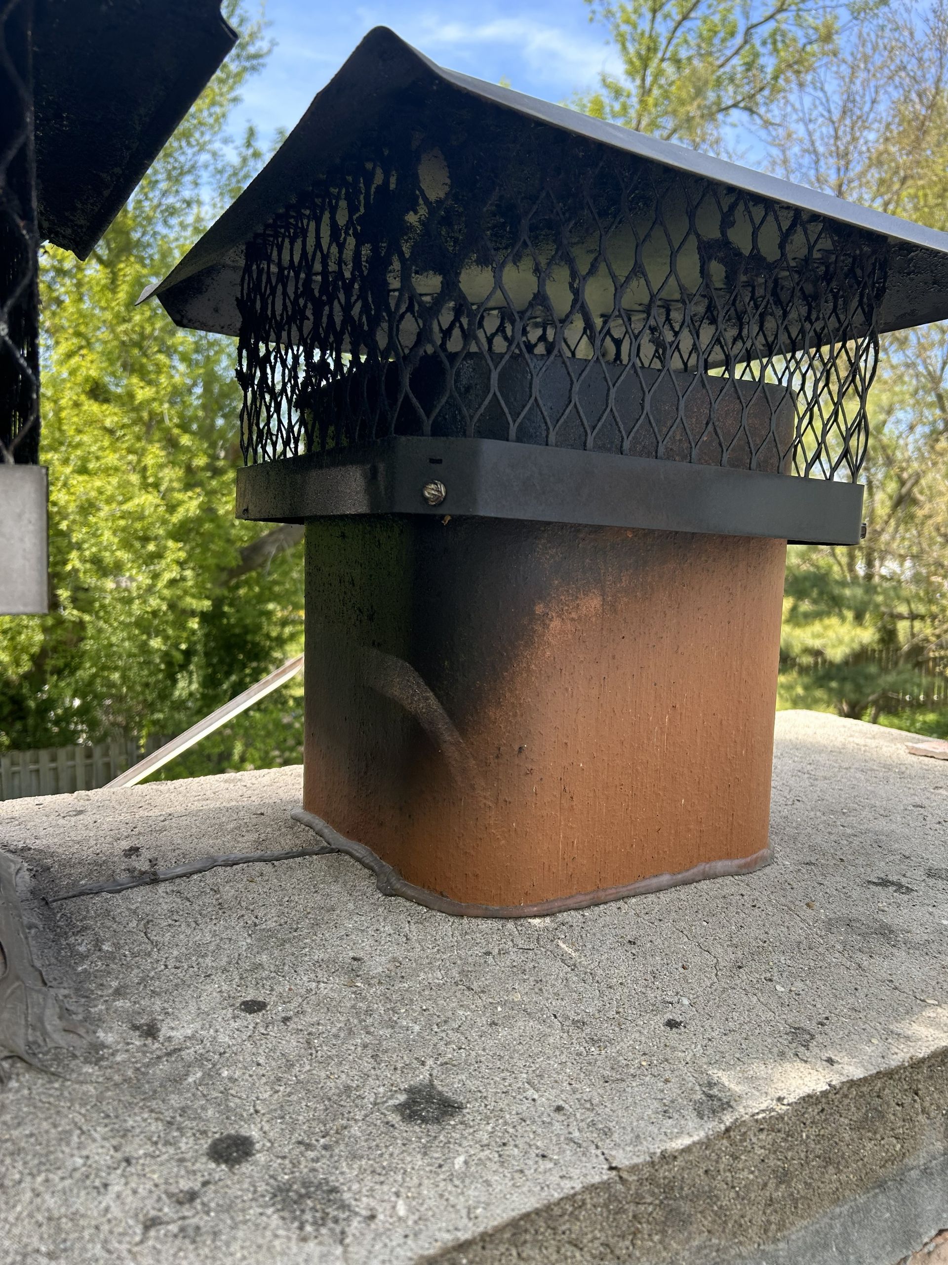 Brick chimney with black cap on a concrete surface, green trees in background.