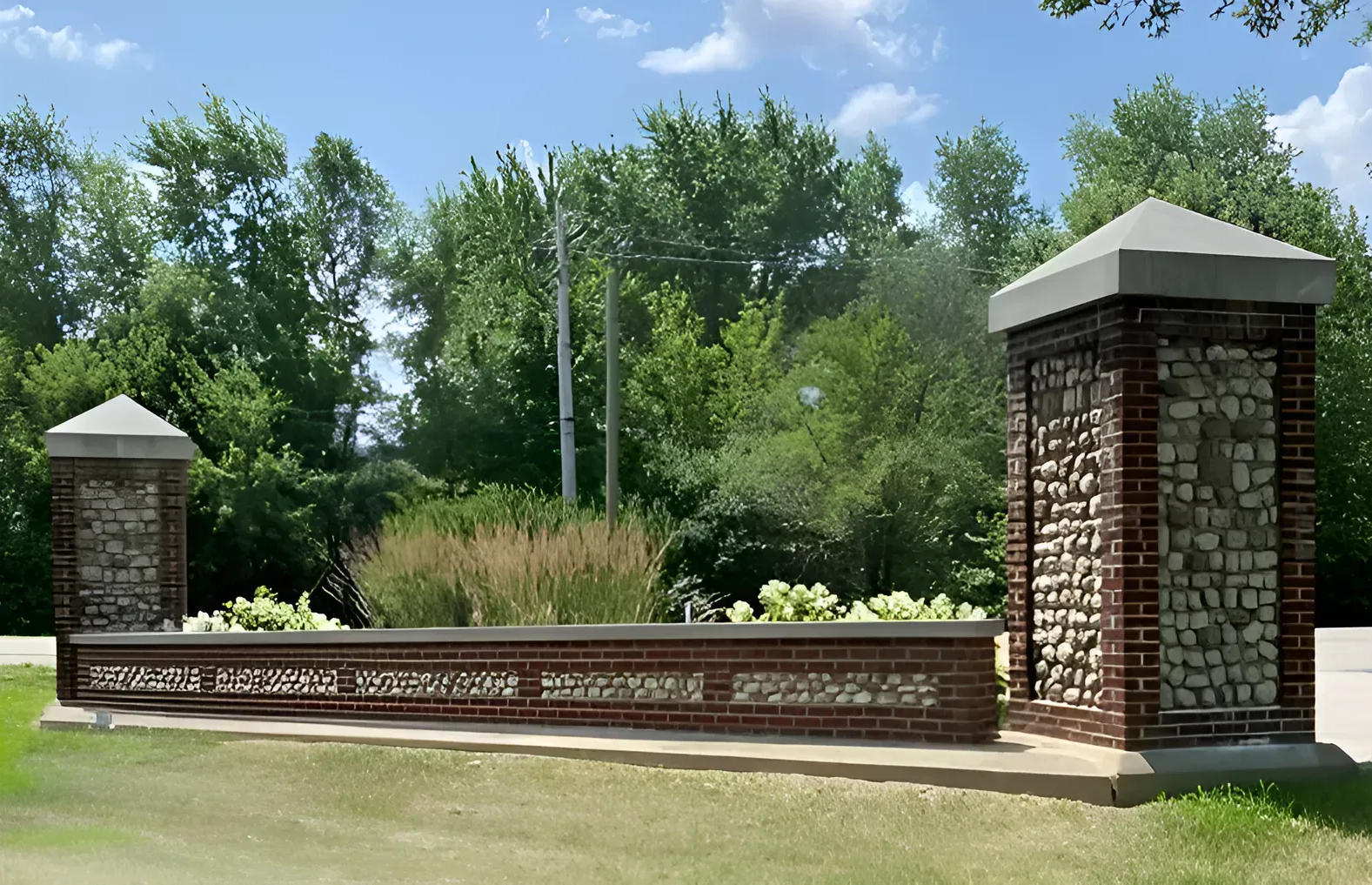 Brick entrance pillars with a low brick wall, flanked by greenery and trees. Blue sky.