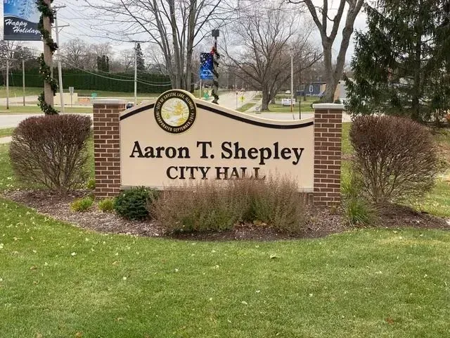Sign for Aaron T. Shepley City Hall, with brick columns and a beige background, in front of bushes and a green lawn.